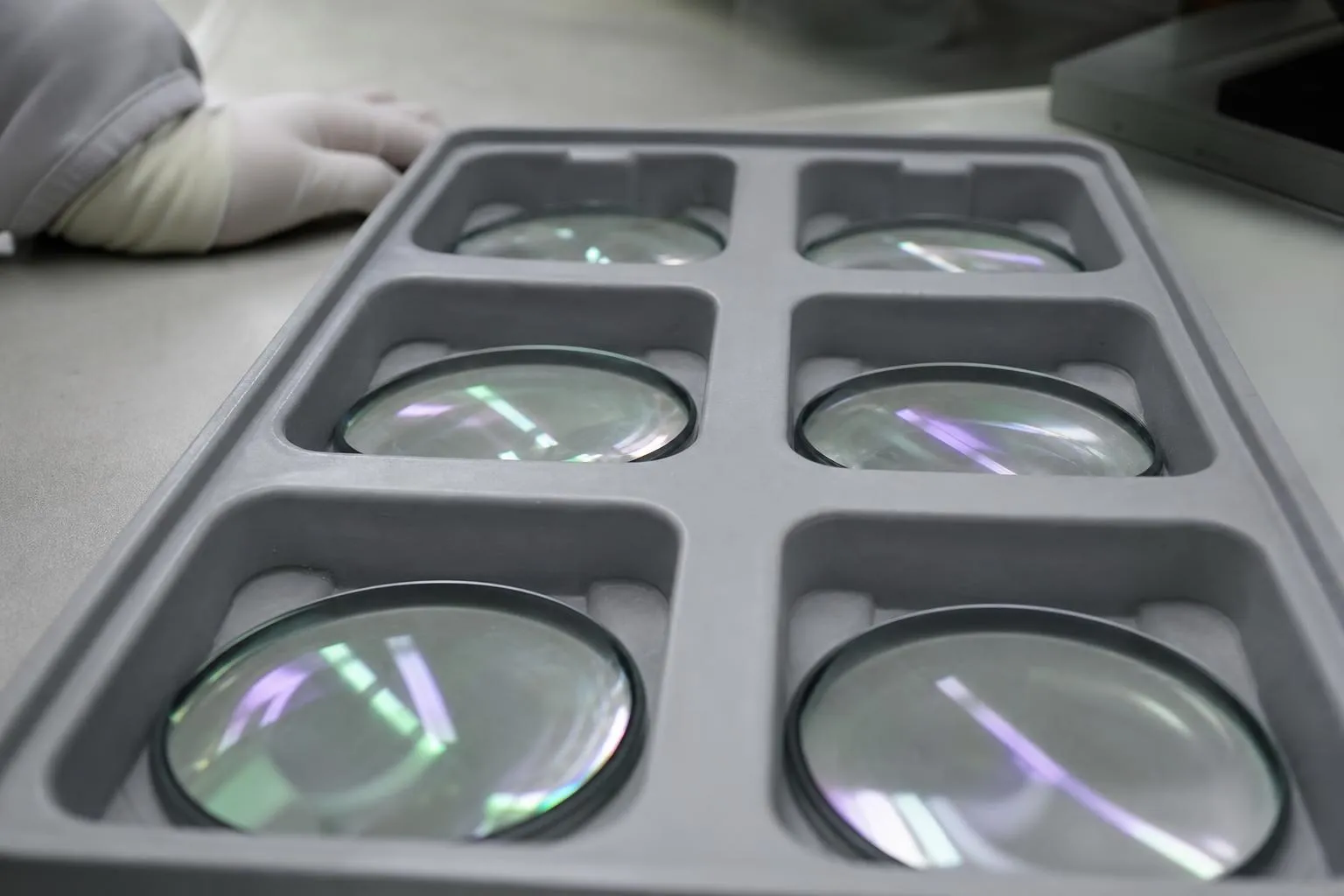 Uncut coated lenses in a grey tray on a lab bench, catching faint green reflections