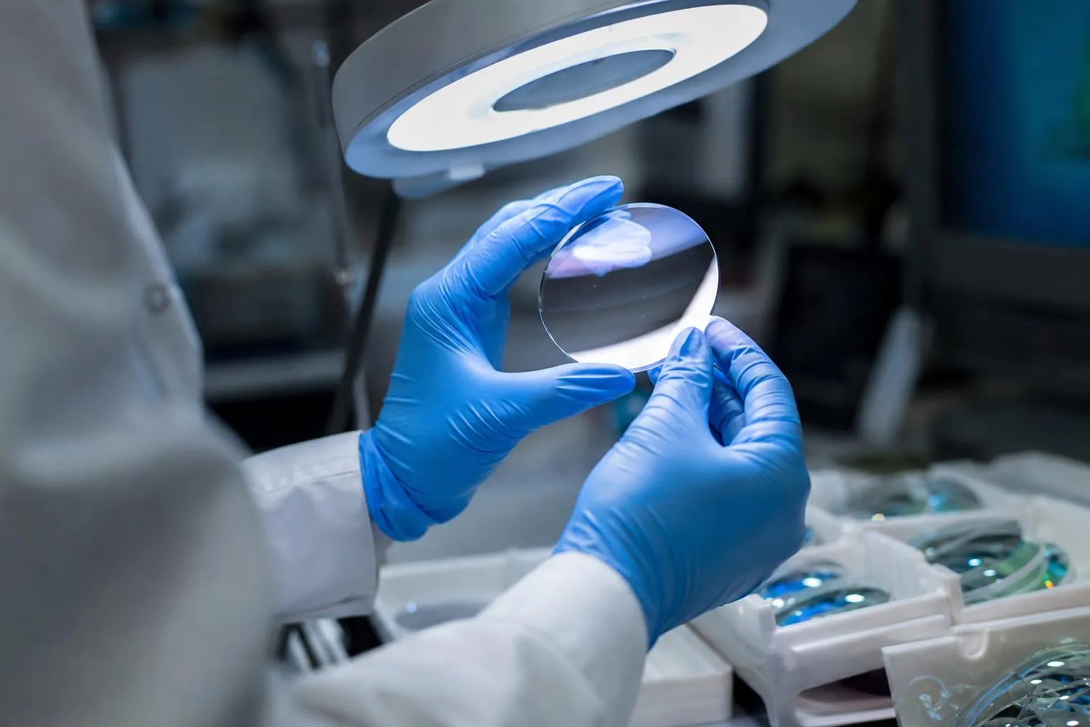 Gloved hands tilting a lens under a bright lamp to inspect its coated surface