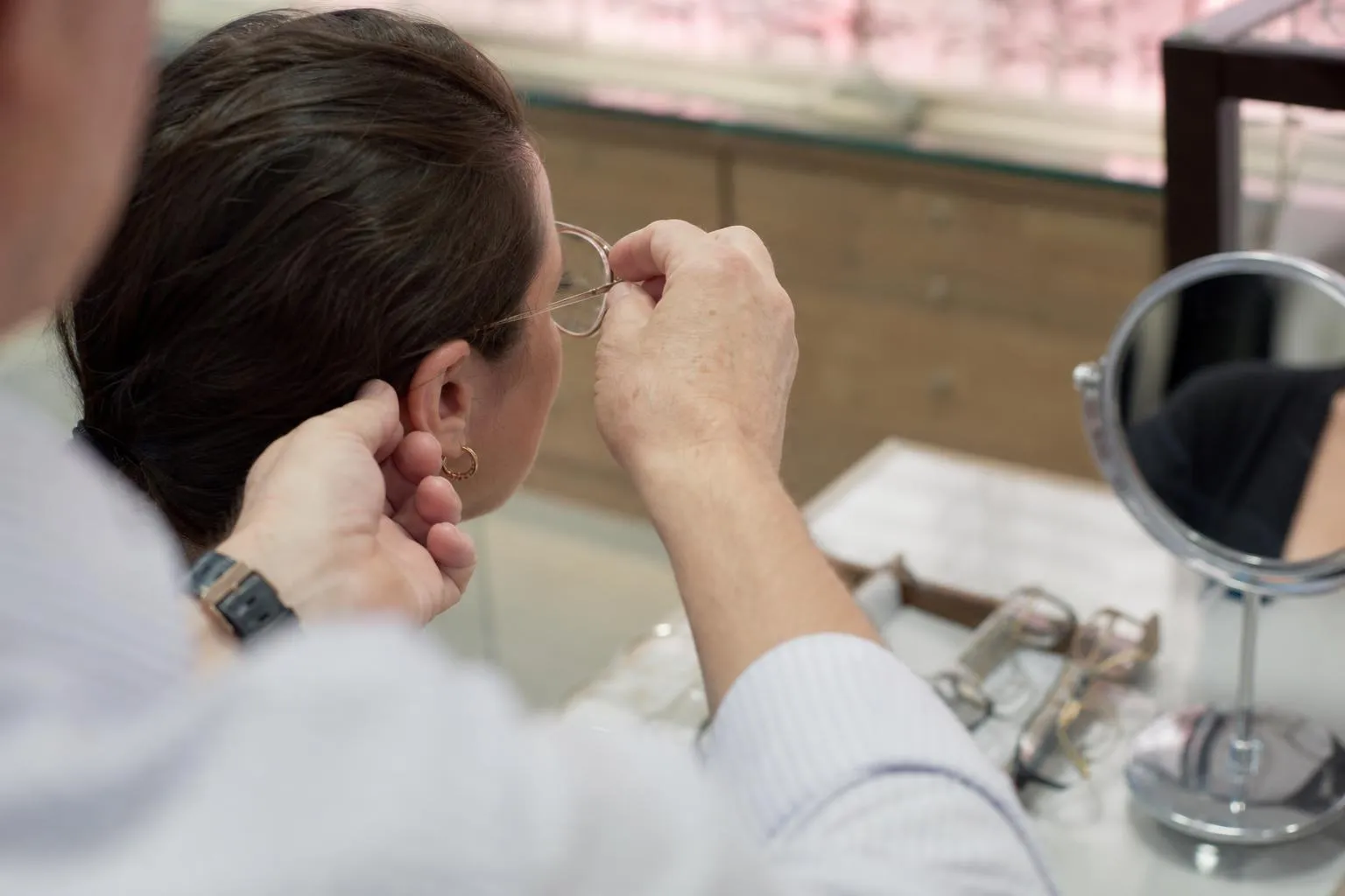 An optician settles a new frame on a seated patient at a dispensing table