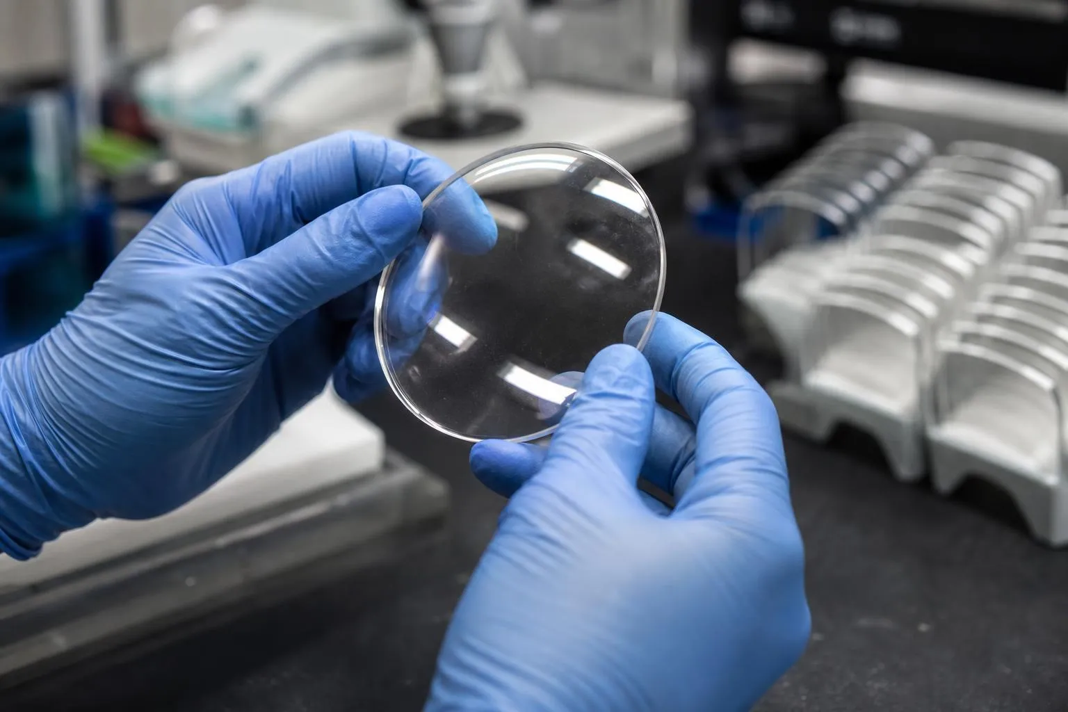 Gloved hands holding a round, uncut lens up to the light at a lab bench