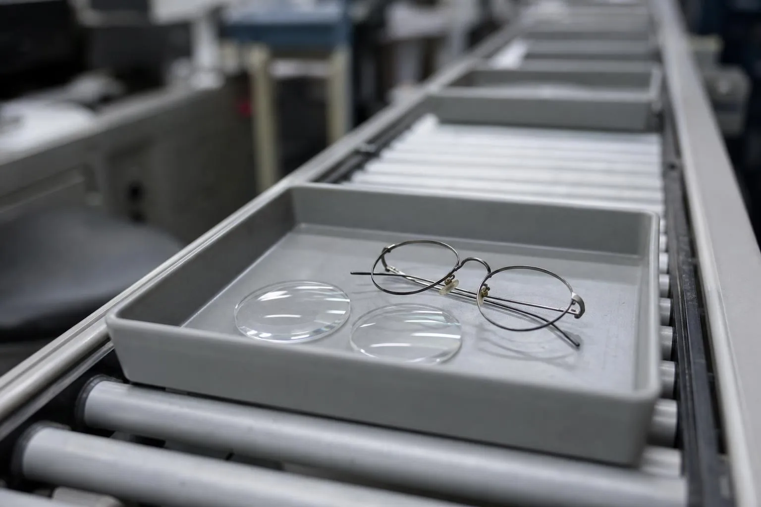 Plastic job tray on a lab conveyor holding two lens blanks and a metal eyeglass frame