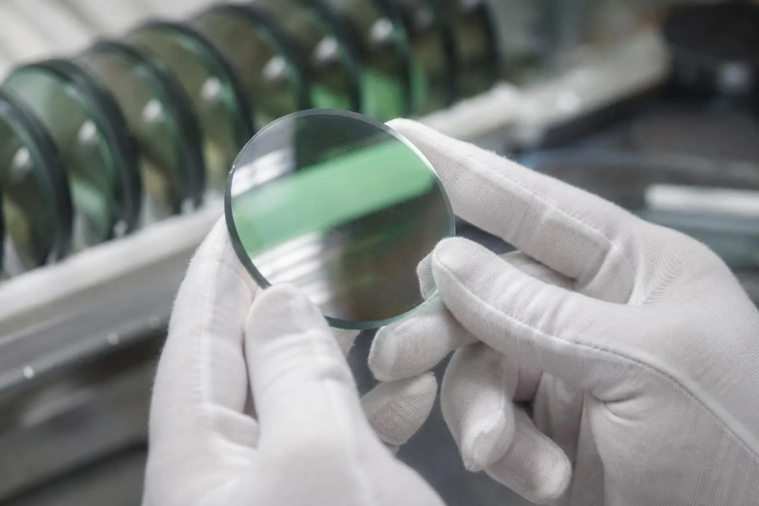 Gloved hands tilting a coated lens under bright light, a faint green reflection on its surface
