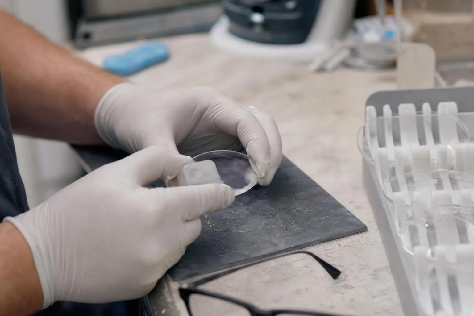 Gloved hands fixing a small block to a round lens at a workbench, with an empty frame and a lens tray beside them