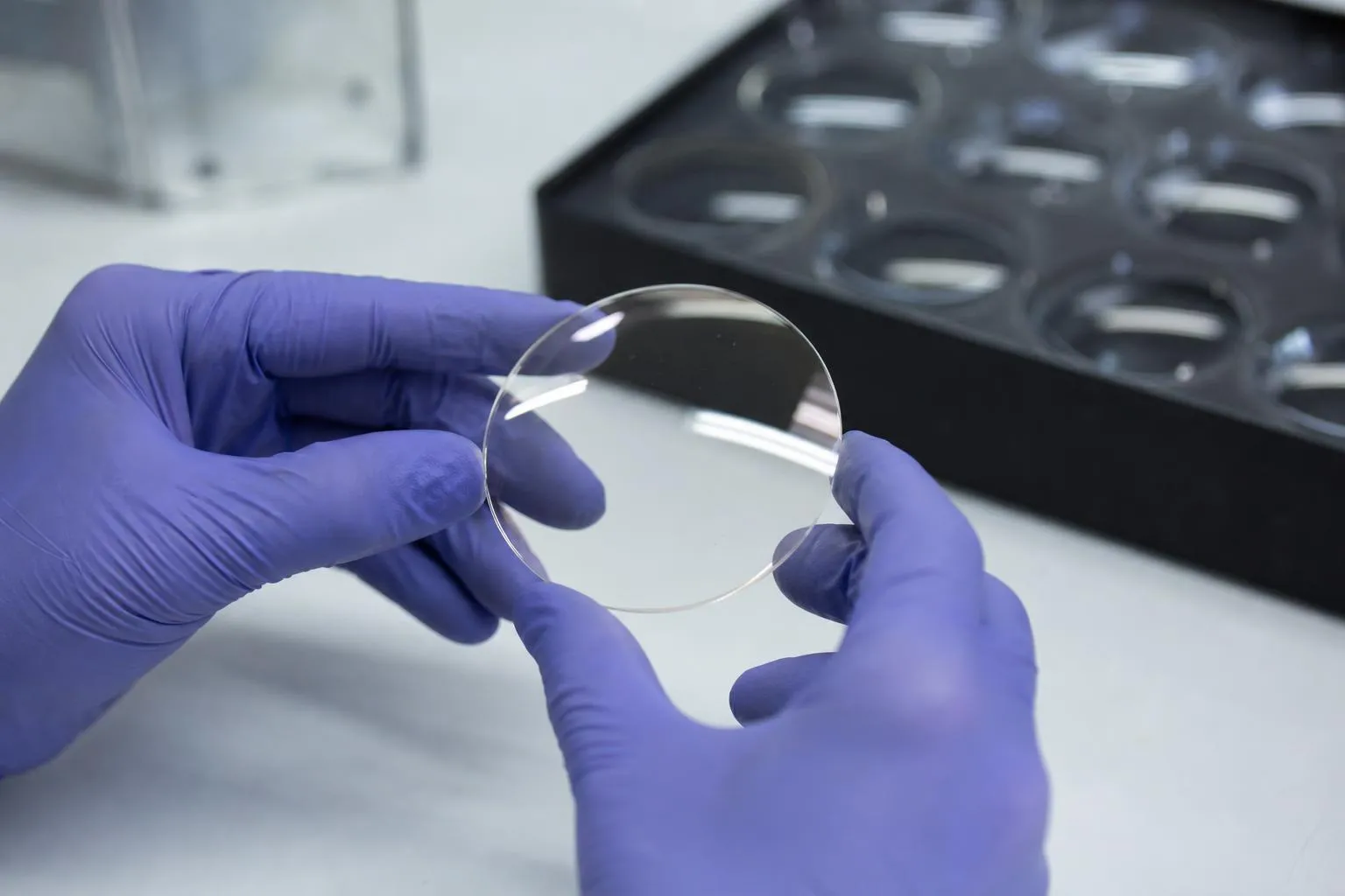 Gloved hands holding a finished clear lens by its edges above a white lab bench