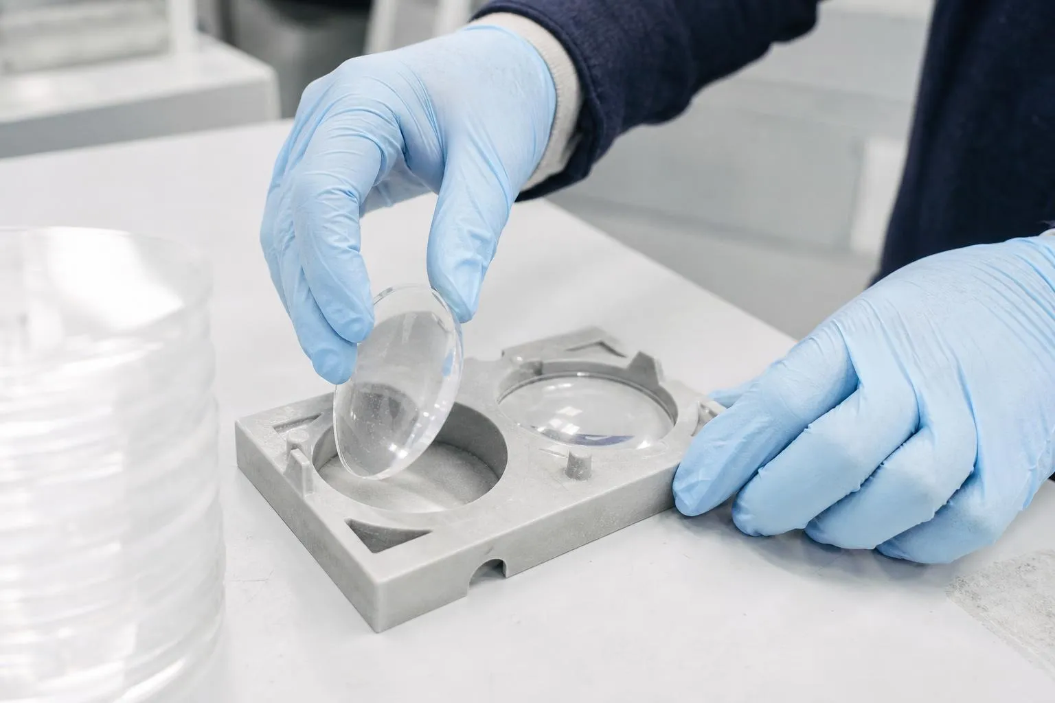 Gloved hands lifting one edged lens from a lens tray on a white lab bench