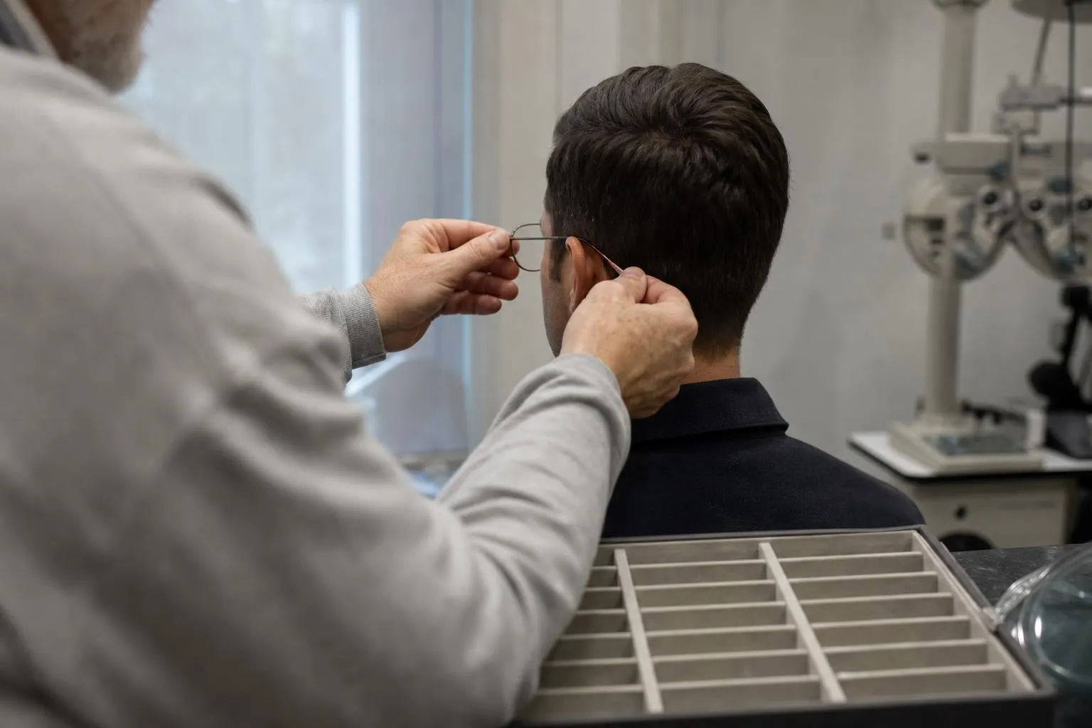 An optician’s hands settling a frame on a seated patient, seen from behind at a dispensing table