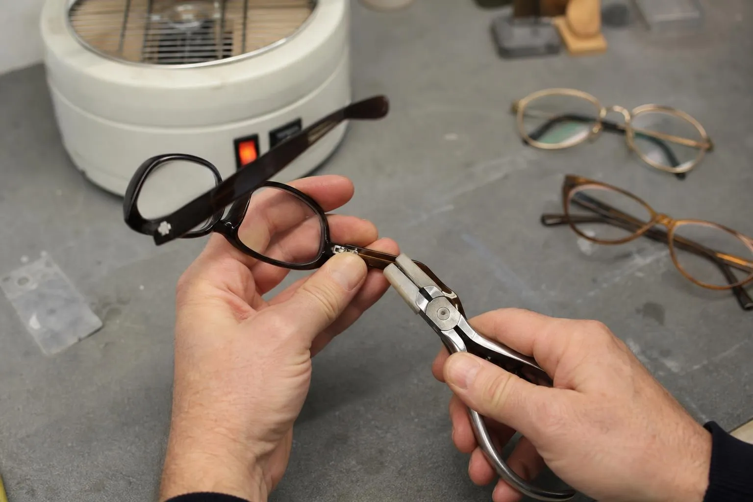 Hands adjusting the temple of an acetate frame with pliers at a dispensing bench